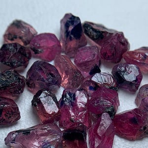 Puede incluir: Una colección de piedras preciosas en bruto, sin cortar, en tonos rosa intenso y púrpura. Las piedras en bruto varían en tamaño y forma, con una apariencia cristalina. Las gemas están dispersas sobre una superficie blanca, mostrando su belleza natural.