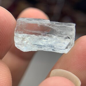 May include: A clear, rectangular gemstone with a slightly rough texture. The gem is held between fingers, showcasing its translucent quality and natural, unpolished appearance. The stone has a light blue hue.