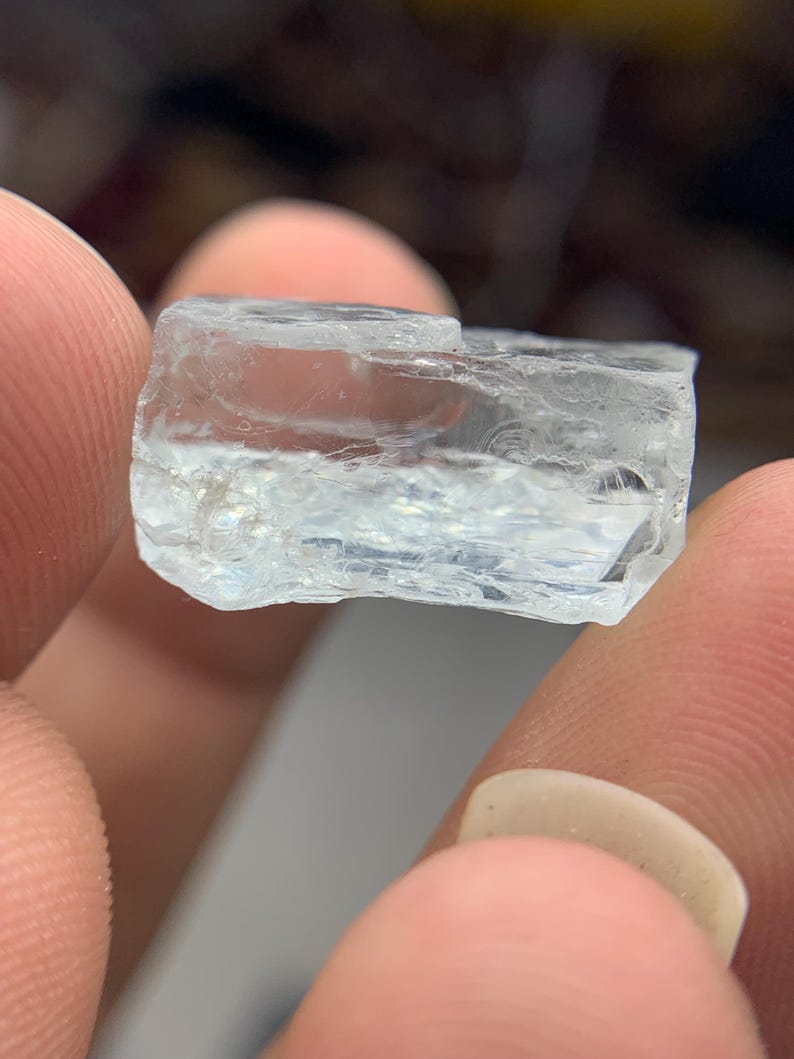 May include: A clear, rectangular gemstone with a slightly rough texture. The gem is held between fingers, showcasing its translucent quality and natural, unpolished appearance. The stone has a light blue hue.