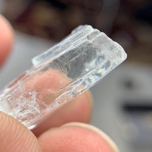 May include: A close-up of a clear, rectangular crystal with a textured surface. The crystal is held between fingers, showcasing its translucent quality and internal fractures. The edges are slightly rough.