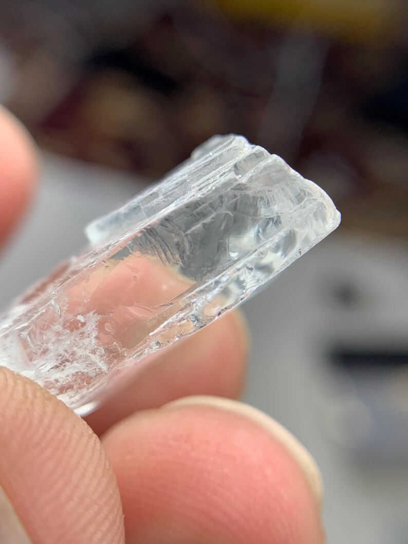 May include: A close-up of a clear, rectangular crystal with a textured surface. The crystal is held between fingers, showcasing its translucent quality and internal fractures. The edges are slightly rough.