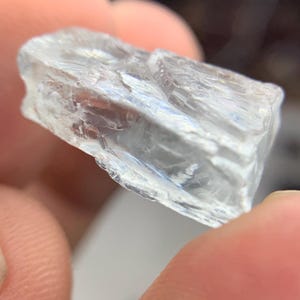 May include: A clear, crystalline mineral specimen with a rough, textured surface. The translucent stone is held between fingers, showcasing its natural, irregular shape and internal structure. The color is a pale, almost colorless, transparent hue.