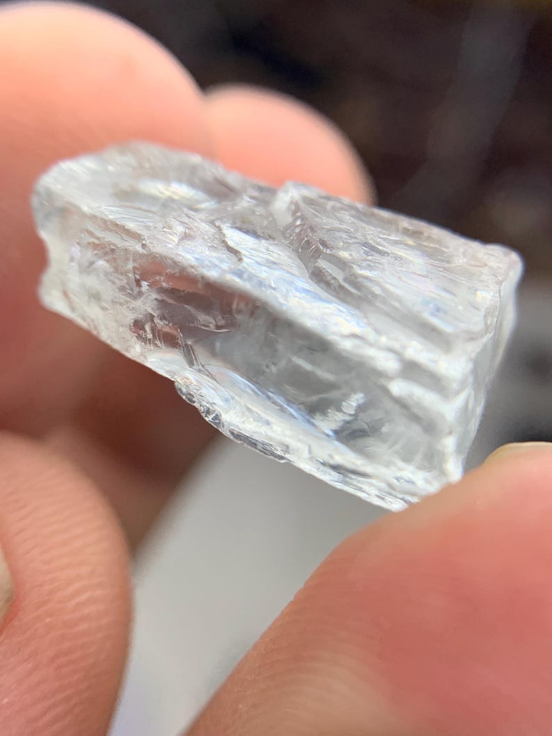 May include: A clear, crystalline mineral specimen with a rough, textured surface. The translucent stone is held between fingers, showcasing its natural, irregular shape and internal structure. The color is a pale, almost colorless, transparent hue.