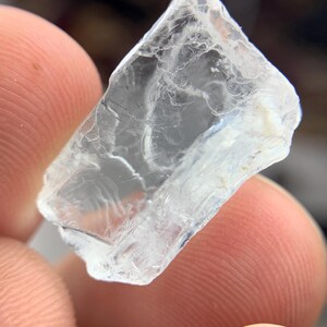 May include: A translucent, crystalline gemstone with a fractured surface. The gem is held between two fingers, highlighting its clarity and natural imperfections. The stone appears to be a rough cut, with sharp edges and a slightly cloudy interior.
