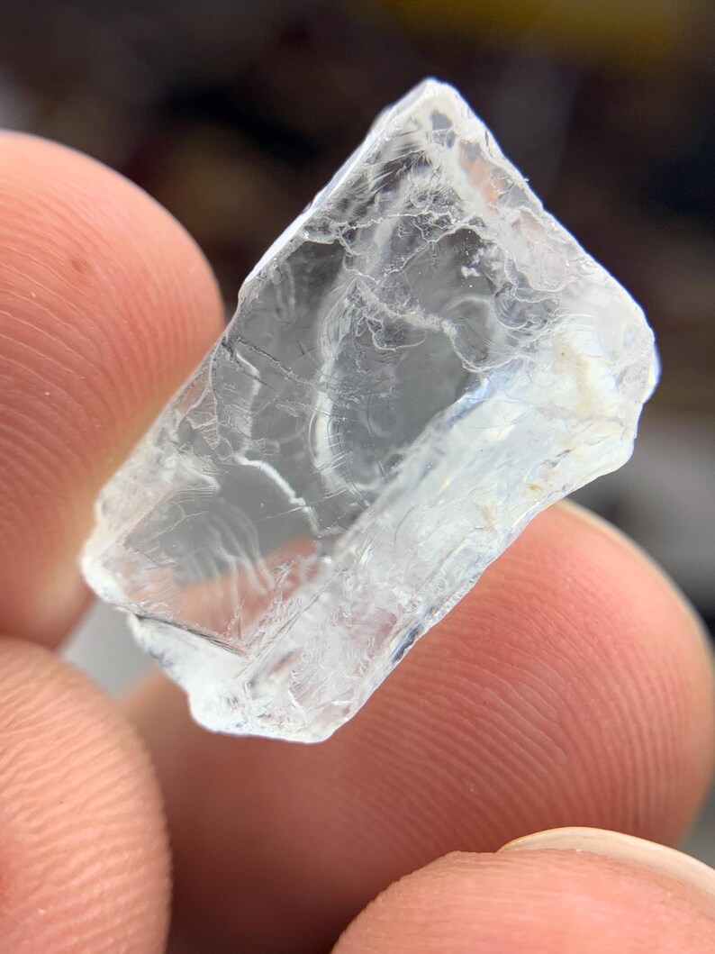 May include: A translucent, crystalline gemstone with a fractured surface. The gem is held between two fingers, highlighting its clarity and natural imperfections. The stone appears to be a rough cut, with sharp edges and a slightly cloudy interior.