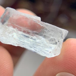 May include: A clear, rectangular crystal with a pale blue tint. The crystal has a rough, textured surface and appears translucent. The edges are sharp and well-defined. The crystal is held between two fingers.