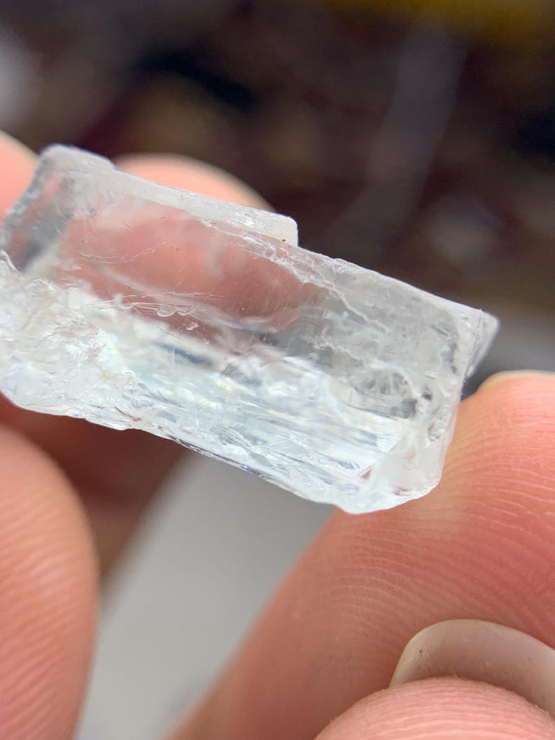 May include: A clear, rectangular crystal with a pale blue tint. The crystal has a rough, textured surface and appears translucent. The edges are sharp and well-defined. The crystal is held between two fingers.