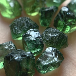 Puede incluir: Una colección de piedras preciosas verdes en bruto, sin cortar. Las piedras varían en tamaño y forma, con una apariencia translúcida y vidriosa. El color varía de un verde claro y brillante a un tono más profundo, similar al de un bosque. Las piedras preciosas están dispersas.