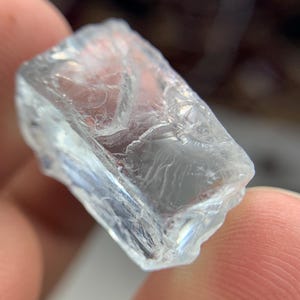 May include: A clear, rectangular crystal with a rough, textured surface. The crystal is transparent, allowing light to pass through, and has a slightly irregular shape. The crystal appears to be a raw, uncut gemstone.