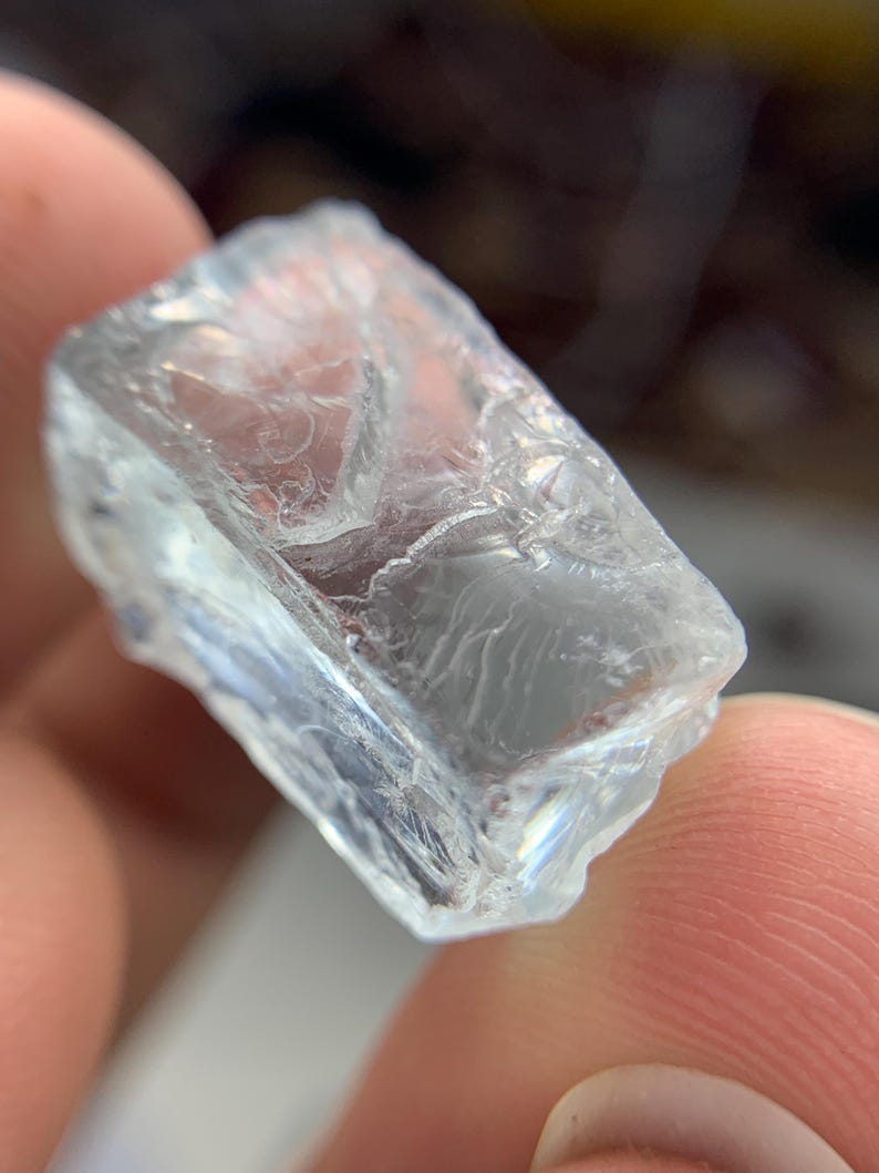 May include: A clear, rectangular crystal with a rough, textured surface. The crystal is transparent, allowing light to pass through, and has a slightly irregular shape. The crystal appears to be a raw, uncut gemstone.