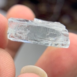 May include: A clear, rectangular crystal with a rough, textured surface. The crystal is held between two fingers, showcasing its translucent quality and natural imperfections. The crystal appears to be a raw, uncut gemstone.