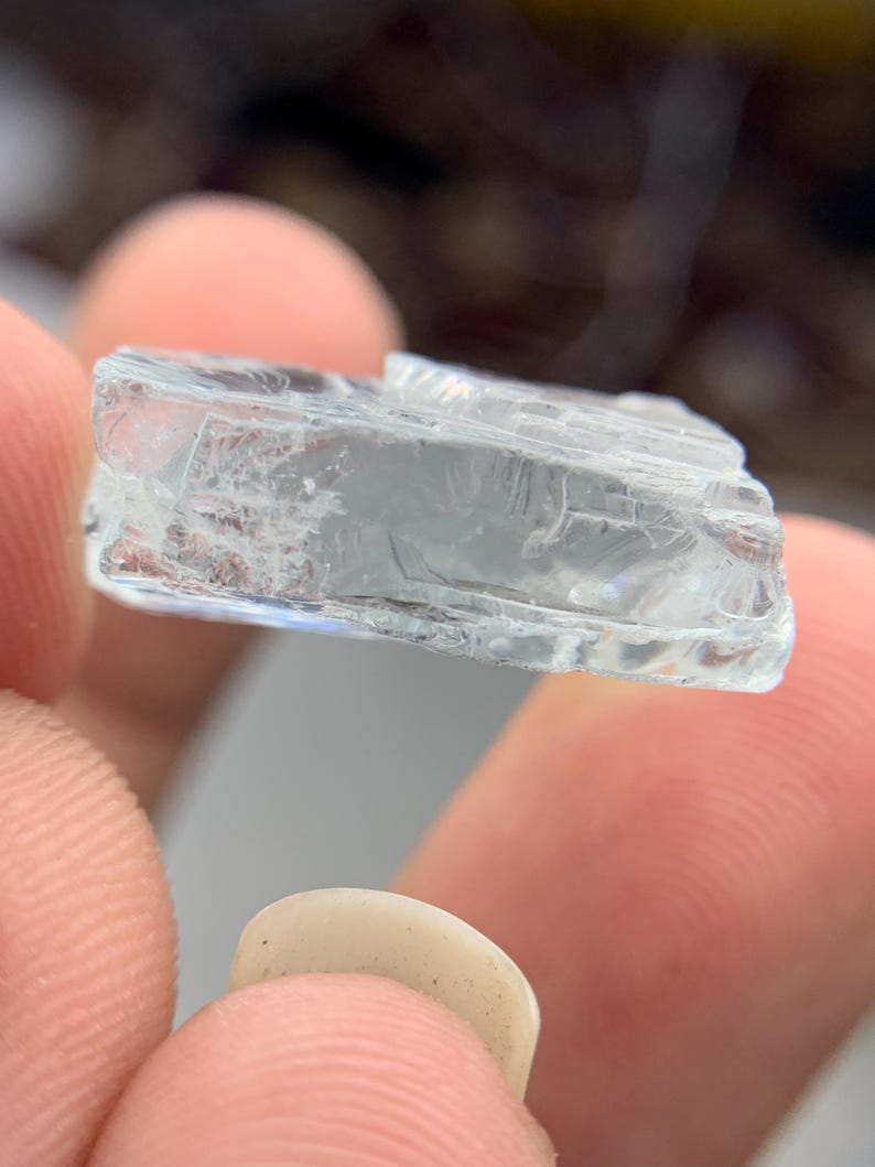 May include: A clear, rectangular crystal with a rough, textured surface. The crystal is held between two fingers, showcasing its translucent quality and natural imperfections. The crystal appears to be a raw, uncut gemstone.