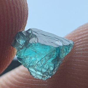 May include: A rough-cut, teal-colored gemstone with a crystalline structure. The gem is held between two fingers, showcasing its natural, irregular shape and translucent quality. The stone's surface reflects light, highlighting its internal facets.