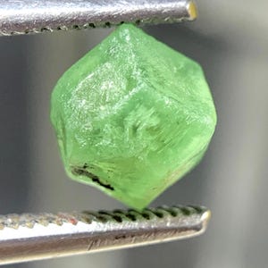 May include: A small, translucent, light green gemstone with a geometric, multi-faceted shape. The crystal displays a vibrant lime-green hue and a natural, raw texture with visible internal inclusions and sharp, angular edges held by metal tweezers.