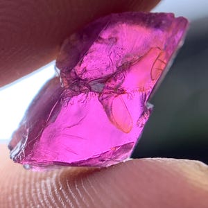 Puede incluir: Una piedra preciosa en bruto y translúcida con un tono magenta rosado vibrante. La piedra presenta una forma irregular y angular con fracturas internas visibles, facetas naturales y una textura brillante y vítrea que refleja la luz.