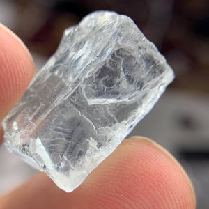 May include: A clear, crystalline mineral specimen with a rough, textured surface. The translucent stone has a fractured appearance, with visible internal structures. The edges are irregular, and the overall shape is roughly rectangular. The stone is held between two fingers.