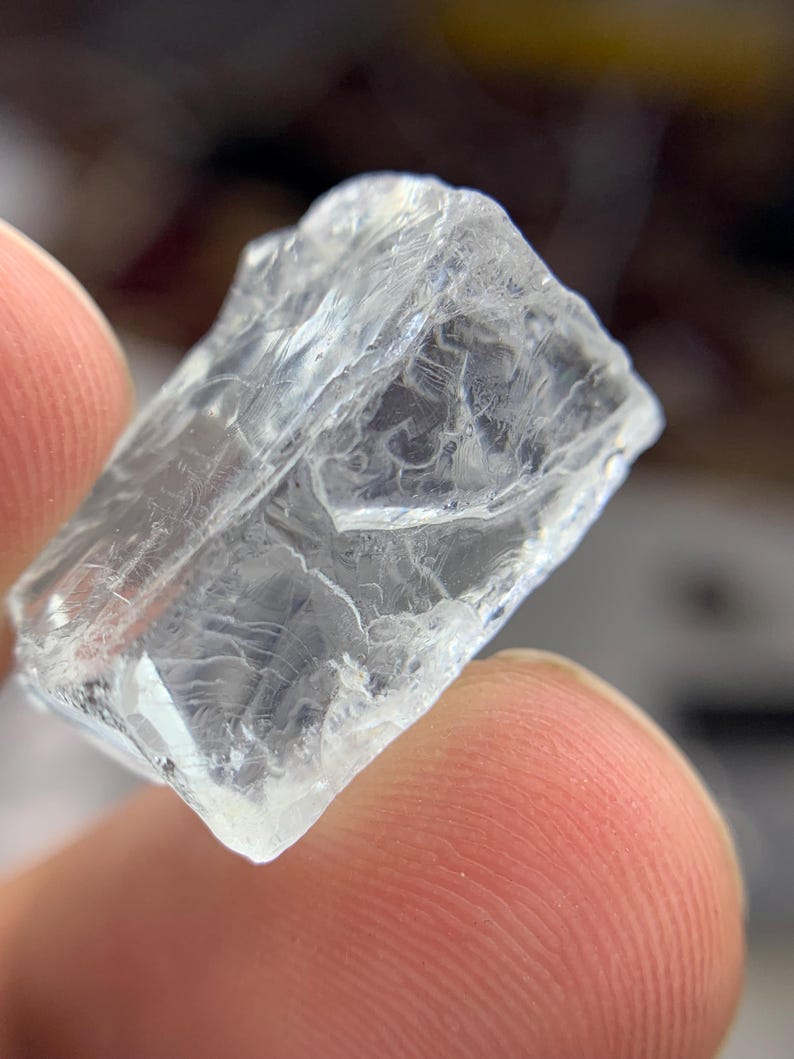 May include: A clear, crystalline mineral specimen with a rough, textured surface. The translucent stone has a fractured appearance, with visible internal structures. The edges are irregular, and the overall shape is roughly rectangular. The stone is held between two fingers.