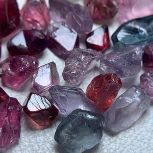 Puede incluir: Una colección de gemas en bruto en varios tonos de rojo, rosa, morado y azul. Las gemas tienen forma irregular y un aspecto natural y sin pulir. La iluminación crea reflejos y sombras en las superficies de las piedras.