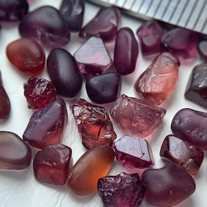 Peut inclure: Une collection de pierres précieuses brutes et polies, dans des tons de violet, de rouge et d'orange. Les pierres varient en forme et en taille, certaines étant translucides et d'autres opaques. Une règle métallique est partiellement visible en arrière-plan.