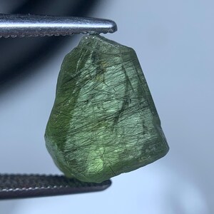 Puede incluir: Una piedra preciosa verde, translúcida y en bruto, con una forma irregular y facetada. La piedra presenta numerosas inclusiones finas de color negro oscuro en forma de aguja en toda su estructura interna, sujeta por pinzas metálicas.