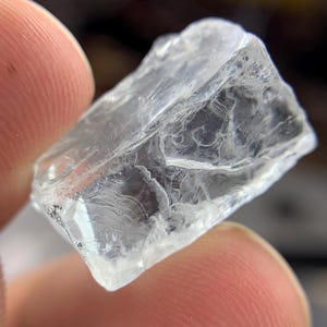 May include: A clear, irregularly shaped crystal with a rough, textured surface. The crystal is held between two fingers, showcasing its translucent quality and internal patterns. The overall appearance suggests a natural, raw gemstone.
