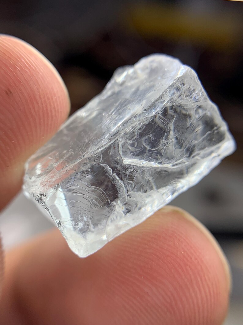 May include: A clear, irregularly shaped crystal with a rough, textured surface. The crystal is held between two fingers, showcasing its translucent quality and internal patterns. The overall appearance suggests a natural, raw gemstone.