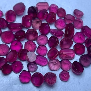 May include: A collection of faceted, deep magenta gemstones. The stones vary in shape and size, with a polished, reflective surface. The gems are scattered across a light-colored surface, creating a visually appealing display of color and light.