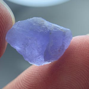 May include: A close-up of a rough, uncut, light purple gemstone. The stone has a textured surface and irregular edges. The gemstone is held between two fingers, with the background out of focus.