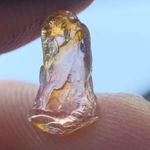 May include: A raw, translucent gemstone with a warm amber and golden-yellow hue. The stone features an irregular, multifaceted surface with natural ridges and indentations, displaying a glassy lustre and internal clarity throughout its structure.