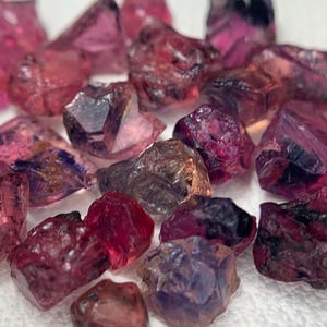 Puede incluir: Una colección de gemas en bruto, sin cortar, en tonos rosa, rojo y púrpura. Las piedras en bruto tienen una estructura cristalina y diferentes niveles de transparencia. Las gemas están dispersas sobre una superficie blanca.