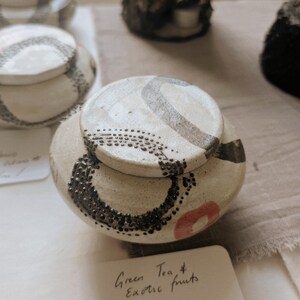 May include: A cream-coloured ceramic jar with a lid, decorated with black dotted and curved patterns, and a red accent. The lid has a grey swirl design. The text "Green Tea & Exotic fruits" is visible on a small card.