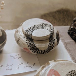 May include: A cream-coloured ceramic jar with a lid, decorated with black and pink patterns. The lid and base have a dotted design. The jar is part of a collection of handmade pottery, ideal for storing small items.