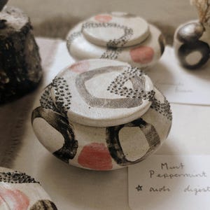 May include: Handmade ceramic lidded jars with abstract black and red designs. The jars have a white base and are decorated with black lines and dots. The lids feature similar patterns. The jars are part of a collection of decorative pottery.