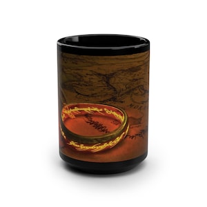 Puede incluir: Taza de cerámica negra con un diseño inspirado en El Señor de los Anillos. El diseño incluye un mapa de fondo y un anillo dorado brillante con inscripciones. La taza es cilíndrica y tiene un interior negro.