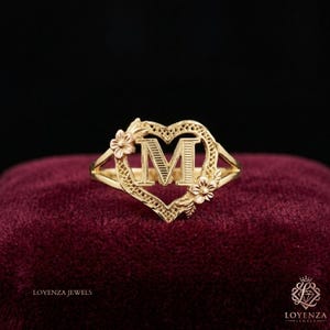 May include: A gold ring featuring a heart-shaped frame with a central letter M. The heart outline has a textured, filigree-style pattern and is accented with two small, detailed floral motifs on the left and right sides of the letter M.