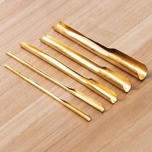 Puede incluir: Una colección de cuatro cucharas y cucharas de té doradas dispuestas en diagonal sobre una superficie de madera marrón claro. Las cucharas tienen un diseño curvo, mientras que las cucharas son delgadas y rectas.