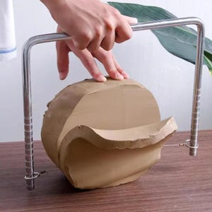 Può includere: Uno strumento di taglio in filo metallico per tagliare l'argilla. Lo strumento è in metallo argentato e ha un filo che attraversa il telaio. Il tagliafilo viene utilizzato per tagliare un grosso pezzo di argilla marrone.