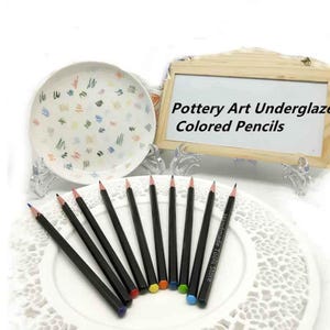 Puede incluir: Una colección de lápices de colores negros con puntas de colores, dispuestos sobre un mantel individual de encaje blanco. Un plato blanco con marcas abstractas coloridas y un letrero de madera que dice "Pottery Art Underglaze Colored Pencils" están en el fondo.