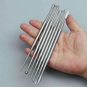 Könnte beinhalten: Eine Sammlung silberfarbener Metallwerkzeuge mit verschiedenen Spitzenformen, darunter spitze, runde und flache Enden. Die Werkzeuge werden in einer Hand vor einem schlichten Hintergrund gehalten. Die Werkzeuge scheinen aus Edelstahl zu sein.