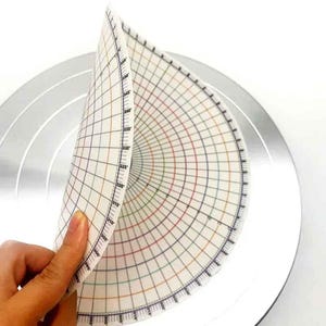 May include: A white and grid-patterned cake decorating stencil is partially lifted, revealing a silver cake stand. The stencil features degree markings and colored lines. The grid is composed of black, red, green, and yellow lines.