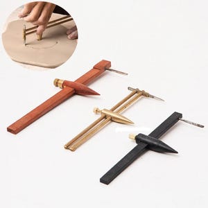 May include: Three pottery compass tools with wooden and metal components. One is dark wood, another is brass, and the third is black. Each tool has a pointed end and a blade for precise clay work. A hand is using a similar tool in the background.