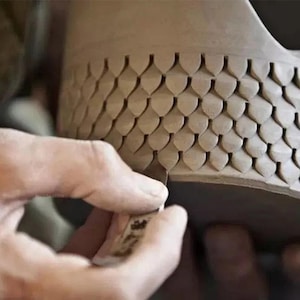 May include: A close-up of a person's hand using a tool to create a pattern of scales on a brown clay pot.