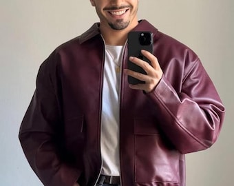 May include: A burgundy faux leather jacket with a zipper closure and two large pockets. The jacket has a ribbed waistband and a relaxed fit. The model is wearing a white t-shirt, blue jeans, and a burgundy baseball cap with a white "NY" logo.
