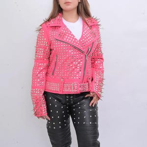 Könnte beinhalten: Eine rosafarbene Lederjacke mit silbernen Nieten und Spikes. Die Jacke hat einen Reißverschluss und einen Gürtel an der Taille.