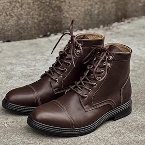 May include: A pair of dark brown ankle boots featuring a cap toe design, matching brown laces, and metallic eyelets. The boots have a smooth finish, a structured silhouette, and a solid black sole with a textured edge detail around the base.
