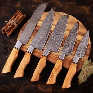May include: A set of five kitchen knives with patterned Damascus steel blades and light brown wooden handles. The knives are arranged on a round wooden cutting board alongside a bundle of cinnamon sticks and a small cluster of dried bay leaves.