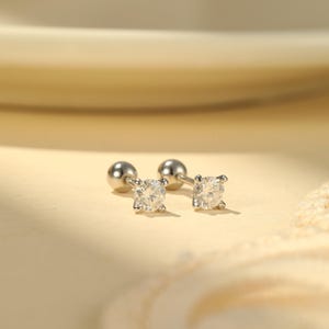 May include: A pair of silver stud earrings with a round, sparkling, clear gemstone. The earrings have a ball-shaped backing. The earrings are displayed on a light beige surface.