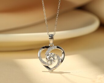 May include: A silver pendant necklace featuring a heart-shaped design. The pendant has a central, round, clear gemstone surrounded by smaller, clear stones. The necklace chain is delicate and silver. The background is a soft, warm color.