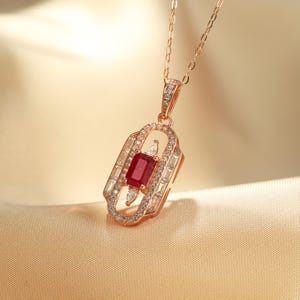 May include: Rose gold necklace with a rectangular ruby pendant, accented with clear, sparkling gemstones. The pendant has an Art Deco design, with a central ruby and surrounding clear stones. The chain is delicate and complements the pendant's elegance.
