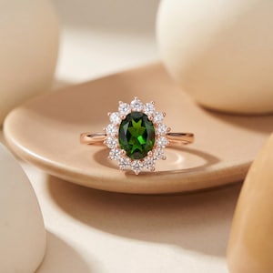 May include: Rose gold ring featuring a large oval-cut green gemstone surrounded by a halo of smaller, clear gemstones. The ring is set on a small, round, light brown plate. The ring's band is thin and delicate.